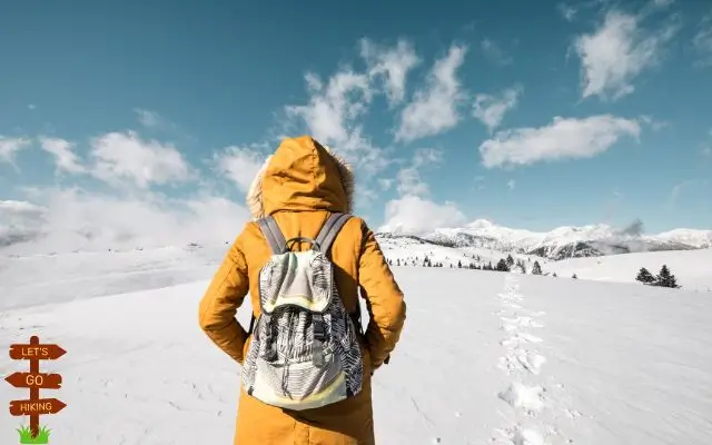 A hiker in a yellow jacket and backpack walking through a snowy landscape, showcasing winter hiking gear against a backdrop of mountains