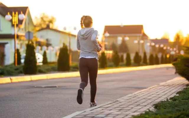 A woman jogging along a quiet suburban street during sunrise or sunset, wearing a hoodie, running shoes, and leggings. 