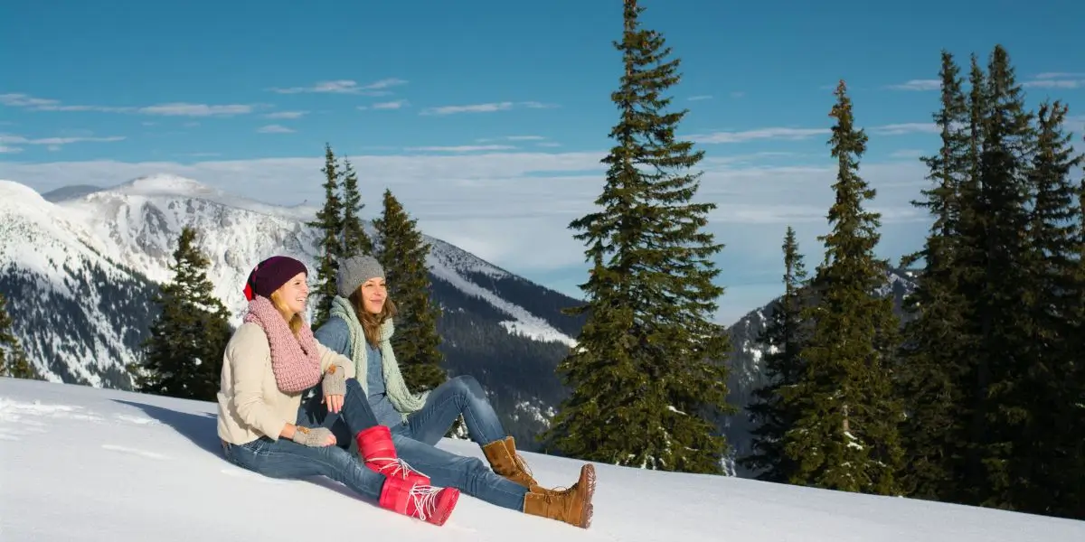 Two women sit on a snowy hillside, dressed warmly in winter clothing, including affordable thermal socks and winter boots.